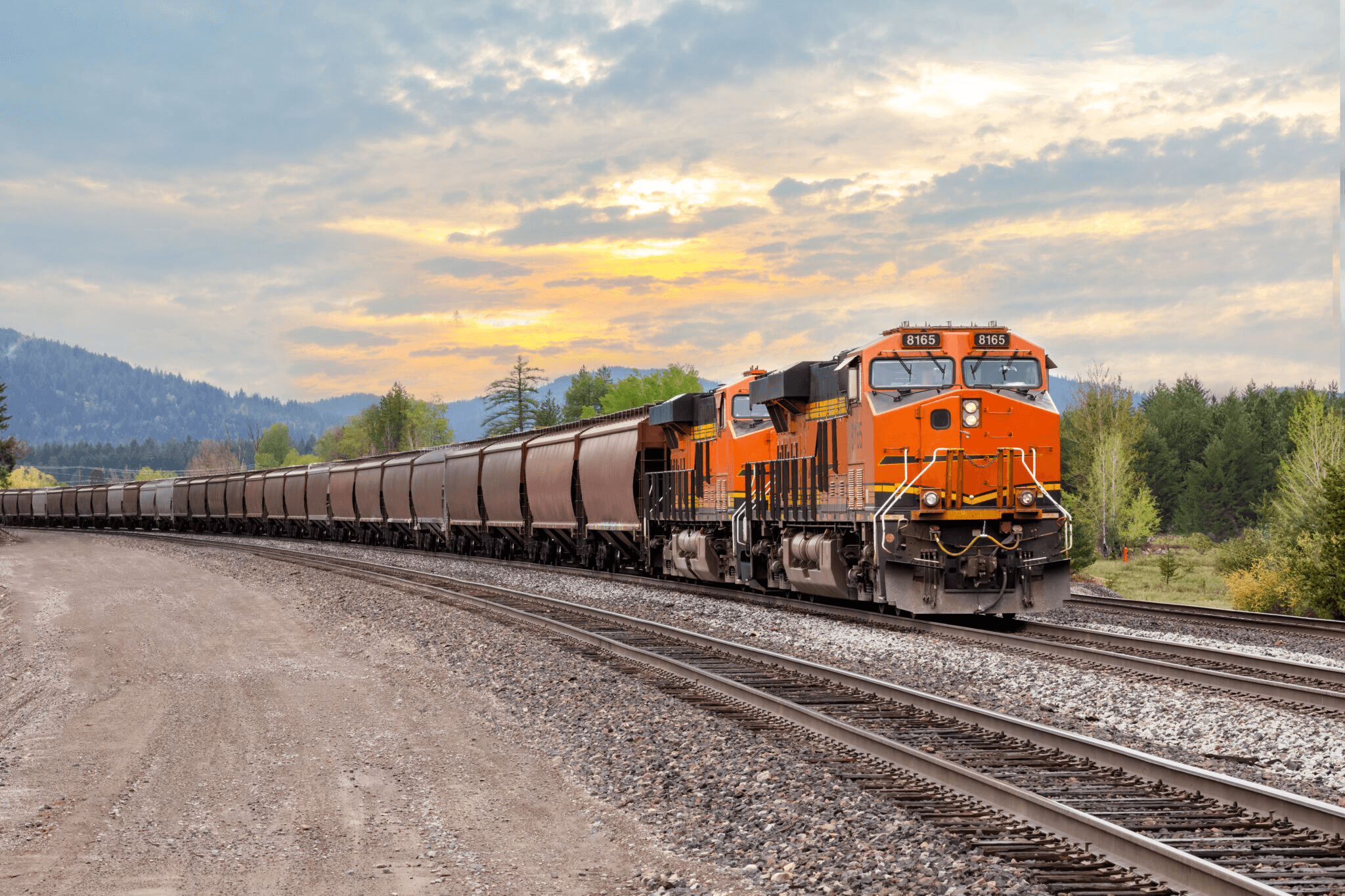 Orange freight train traveling through a forested landscape at sunset.
