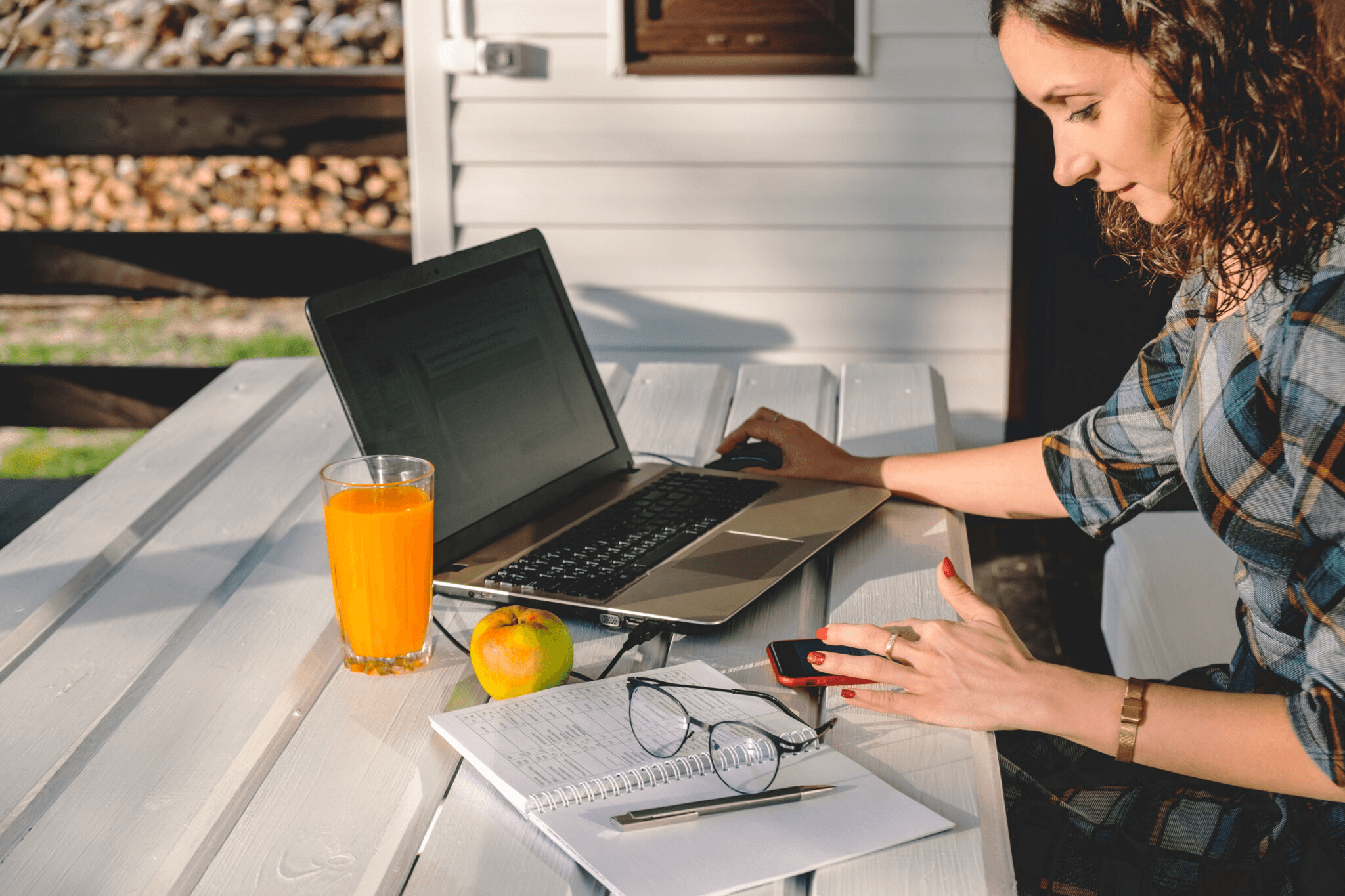 Woman sitting at a desk outdoors with a laptop, phone, and journal.