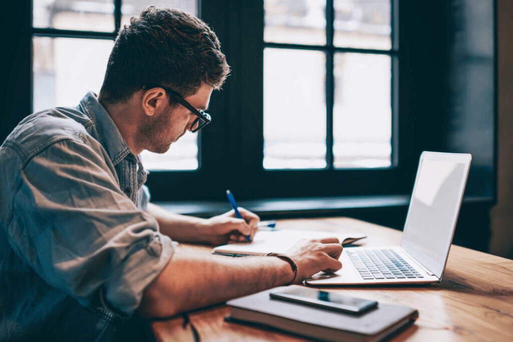 Person working at a desk, writing in a notebook beside an open laptop near a window.