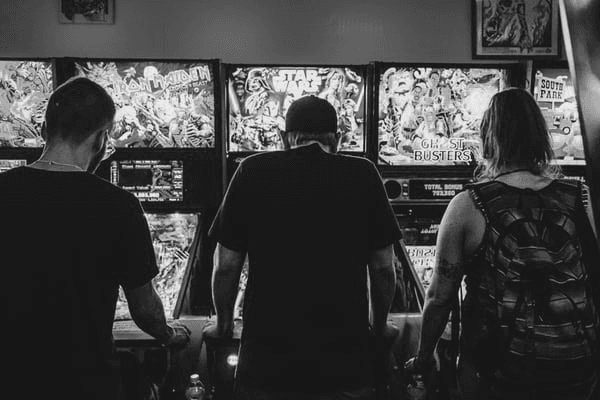 Three people playing pinball machines in an arcade.