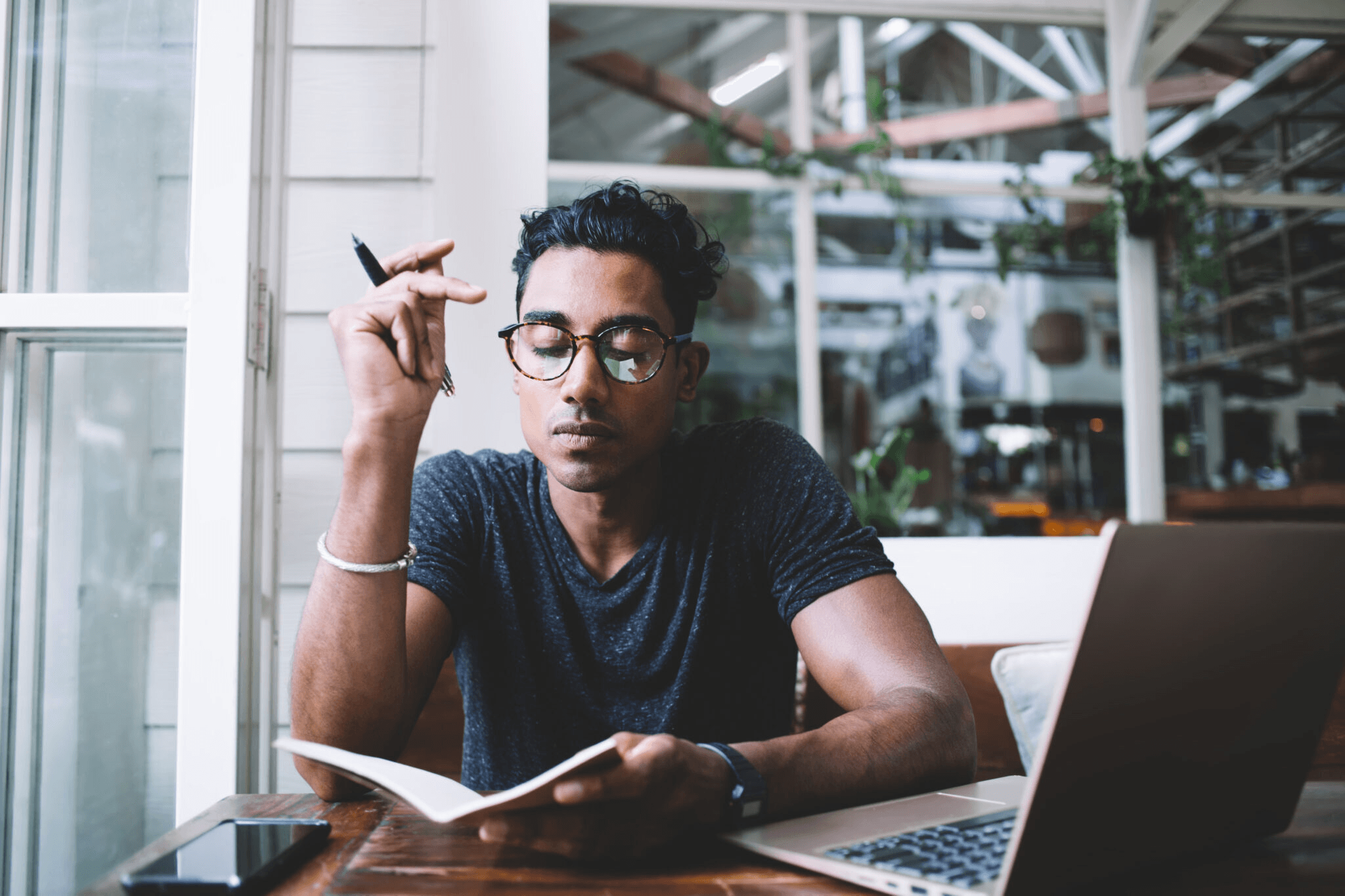 Young man with glasses at a cafe table, writing in a notebook beside a laptop and phone.